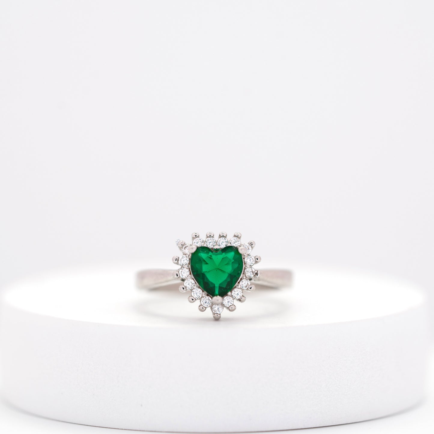 front view of heart emerald silver ring surrounded by diamond clusters placed on a white stand