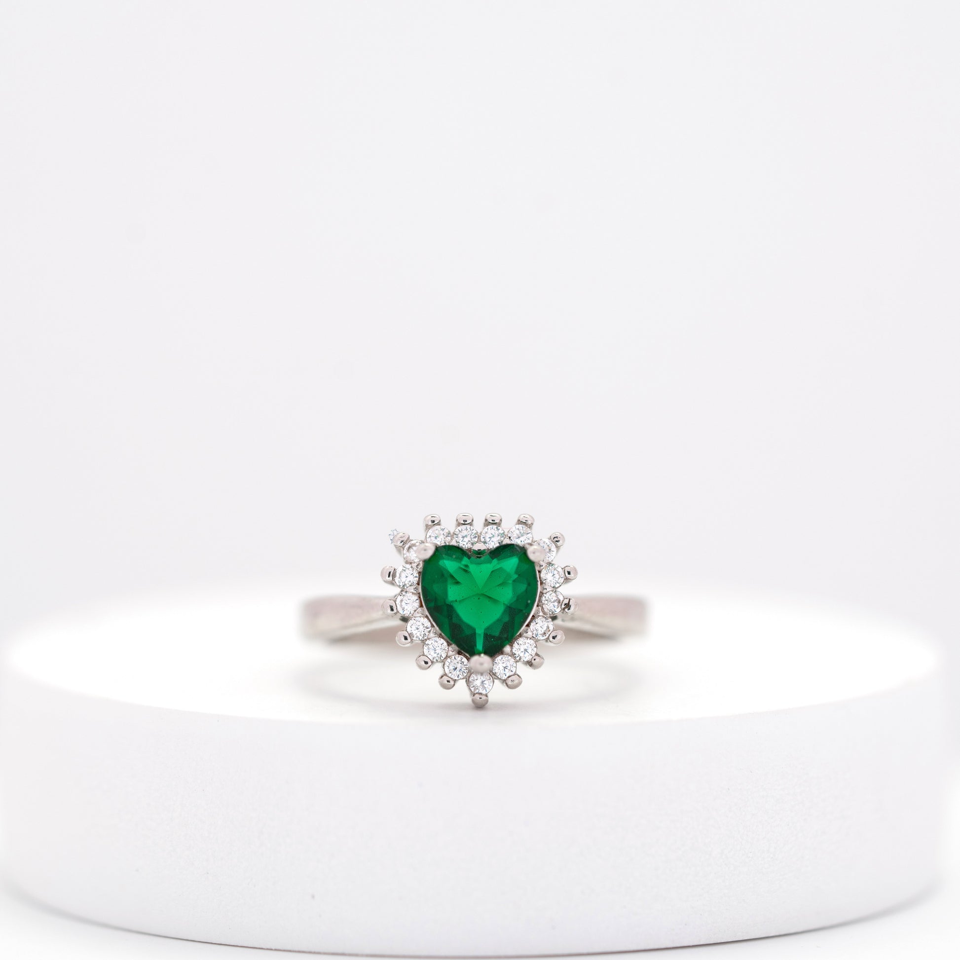front view of heart emerald silver ring  surrounded by diamond clusters placed on a white stand