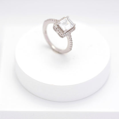 Elegant silver emerald cut ring with halo and pavé setting on a white stand